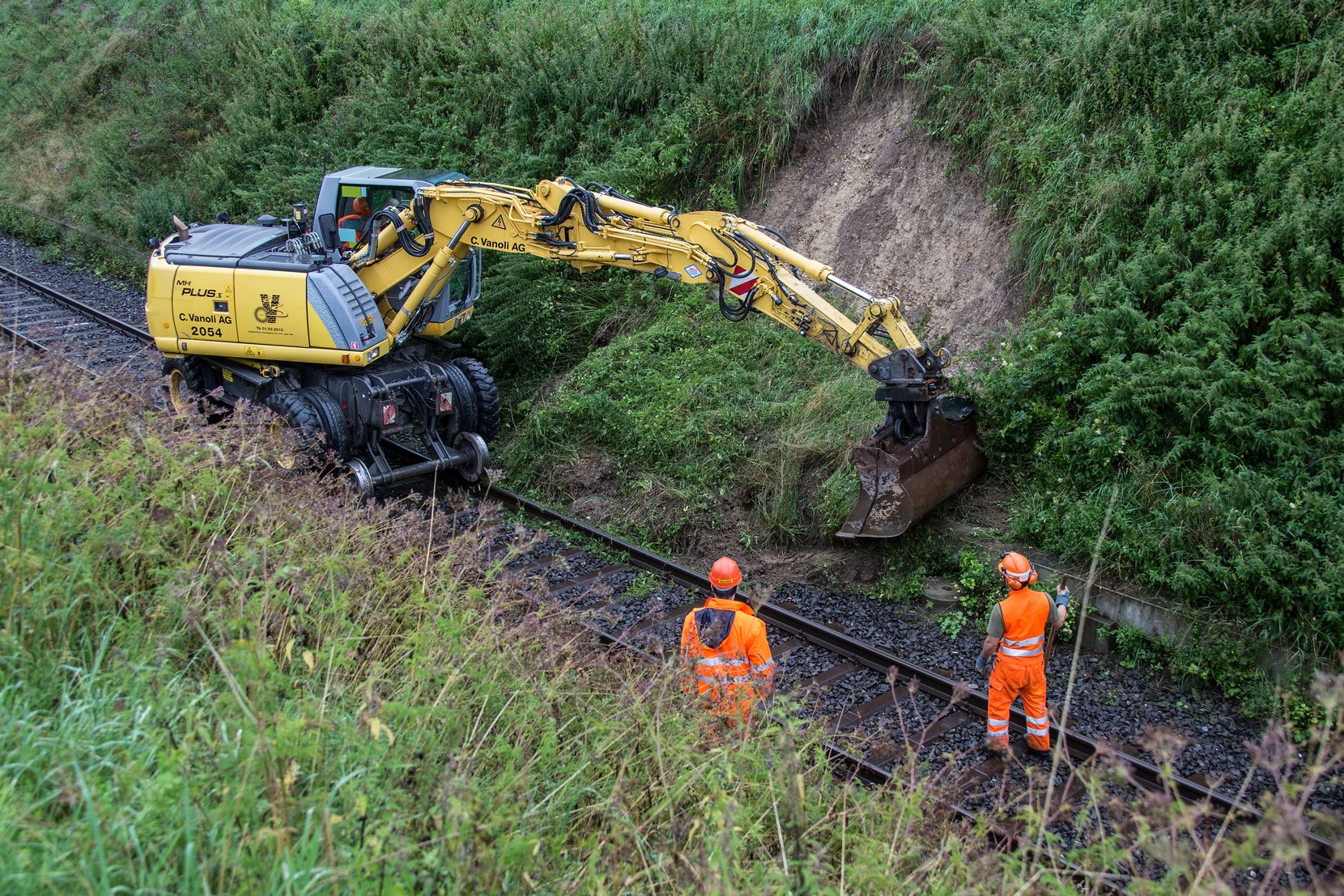 Ein Schienenbagger räumt einen Erdrutsch von den Schienen im Weiler Fistel weg. Er wird dabei von zwei Arbeitern beobachtet.